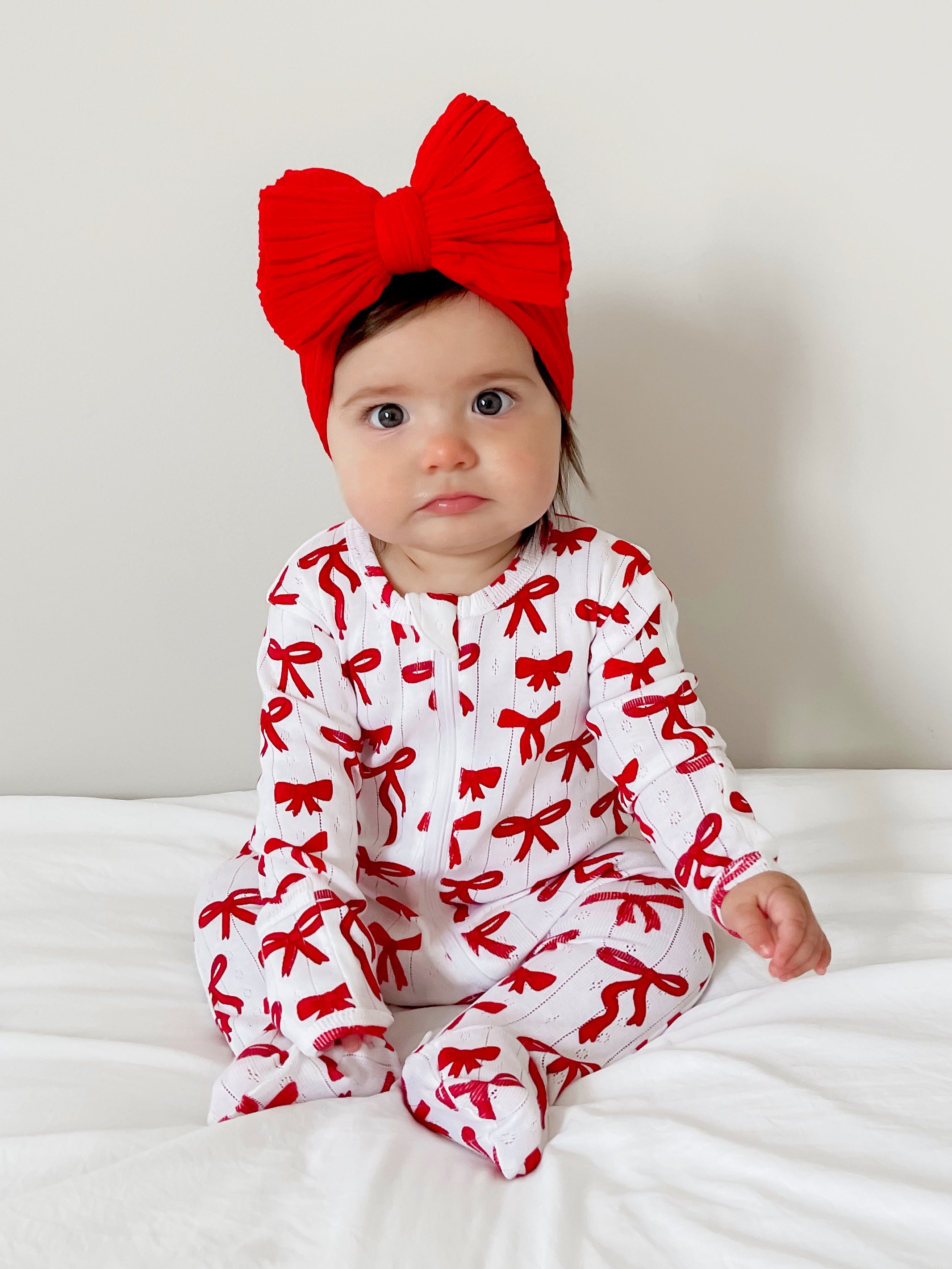 Infant girl in a white outfit with red bows, wearing a large red bow headband, seated on a white bed.