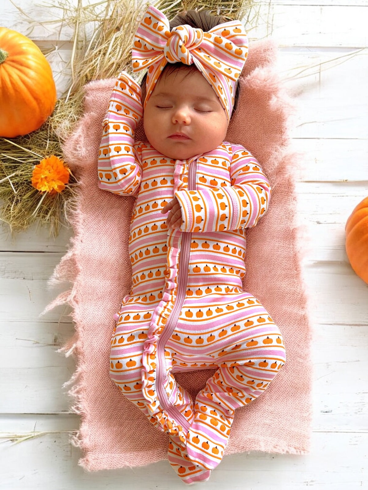 Baby girl in pumpkin-patterned onesie and headband, sleeping on soft pink blanket with pumpkins and flowers around.