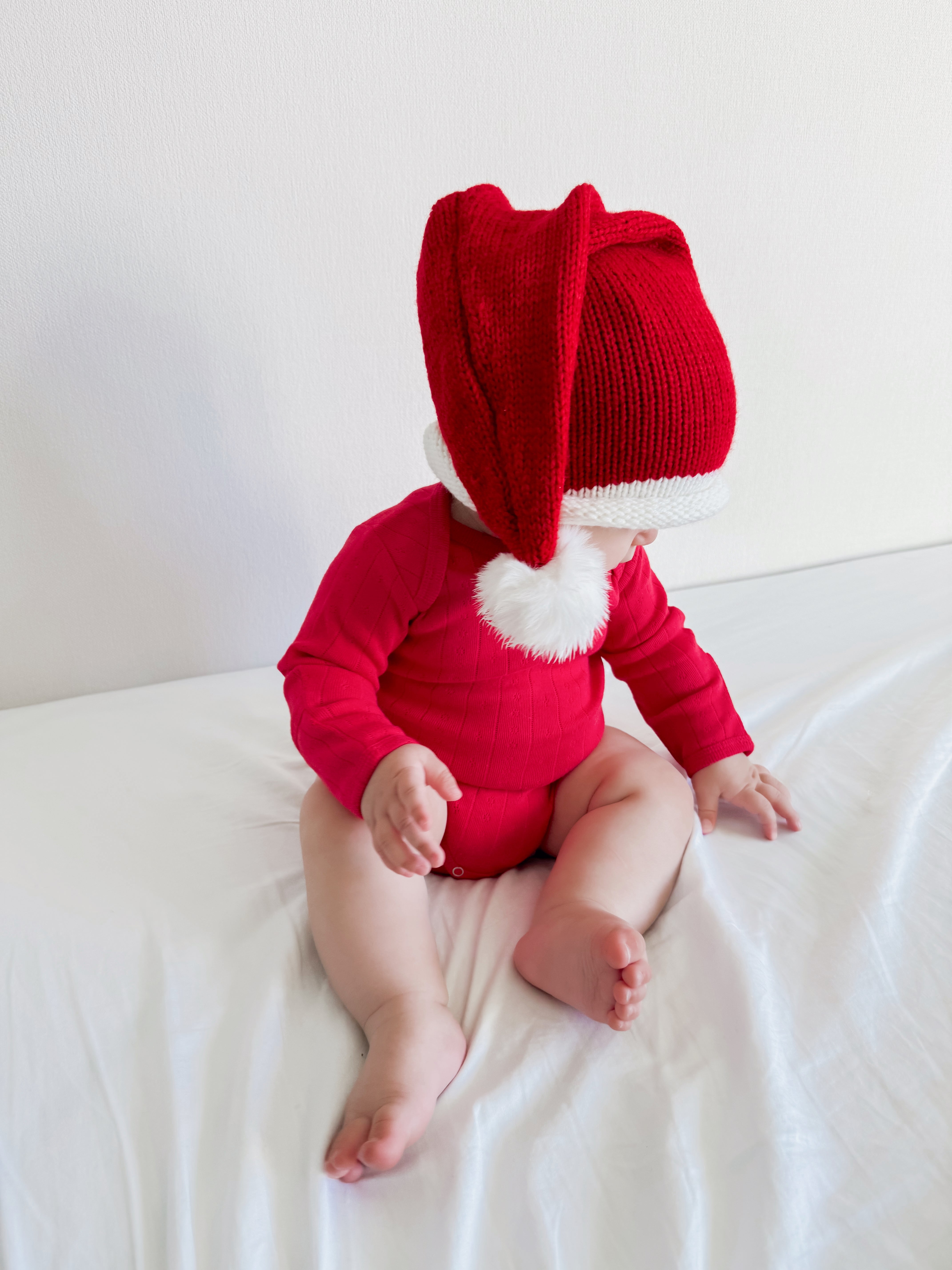 Baby wearing a red holiday outfit and oversized Santa hat, seated on a bed with white sheets.