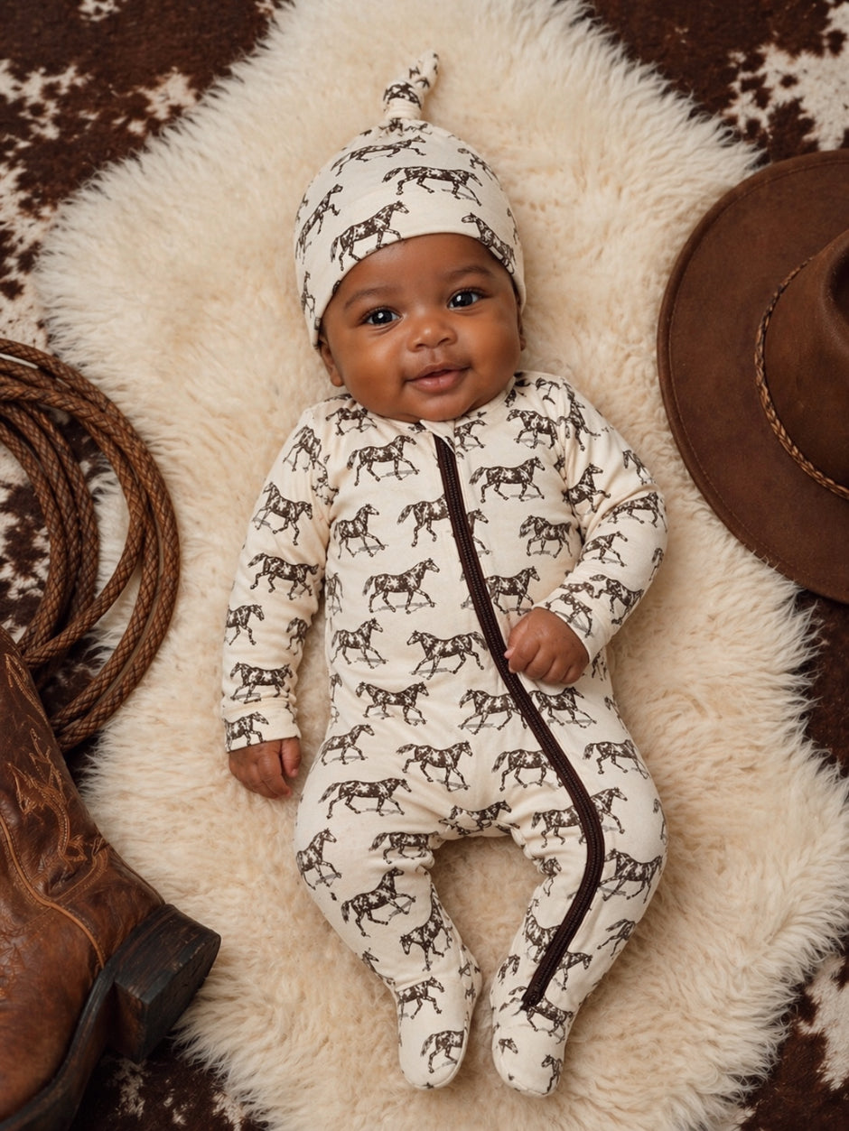 Smiling baby in horse-patterned outfit on soft fur with cowboy hat and boots nearby.