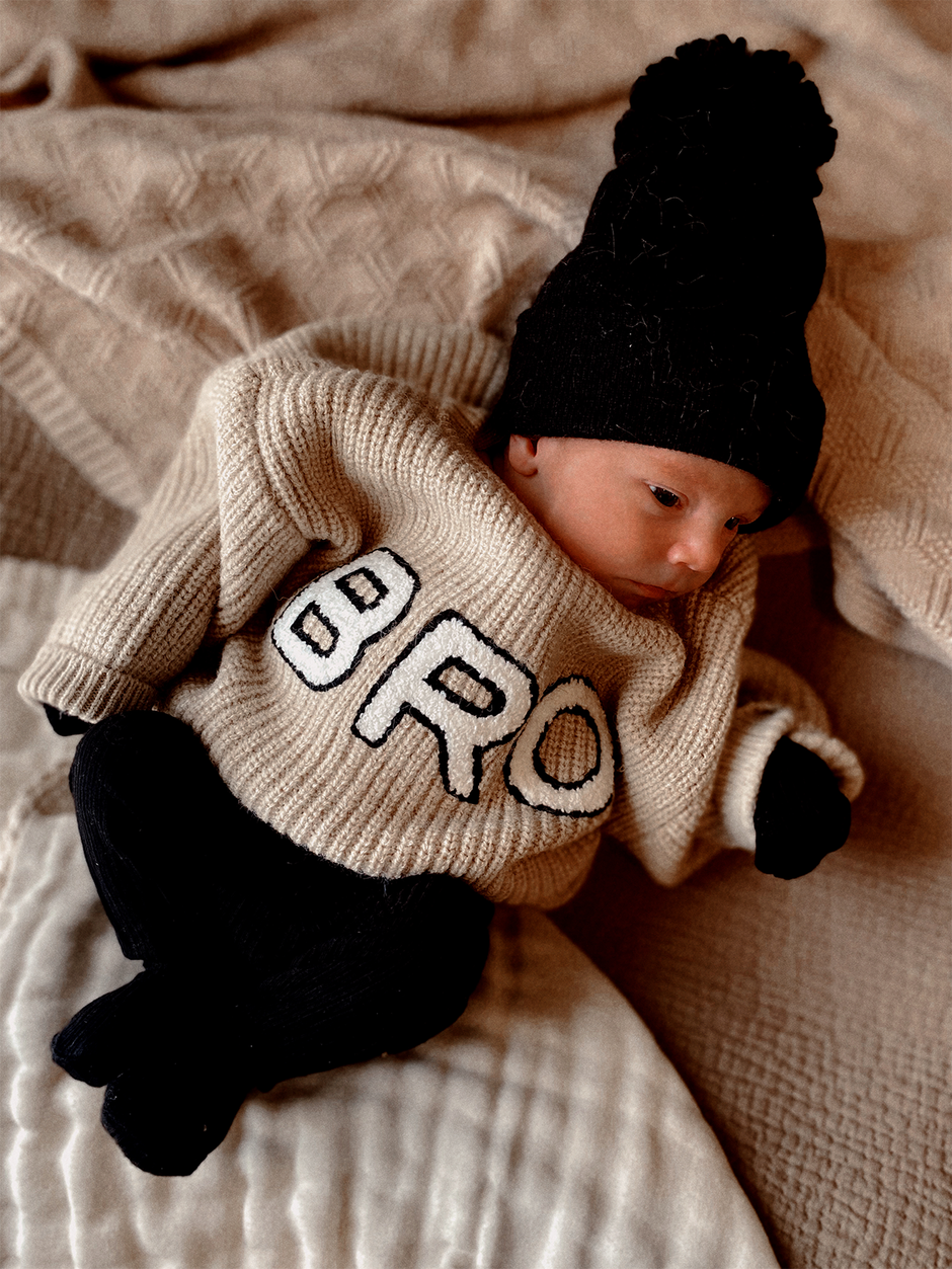 Newborn in a cozy beige sweater with "BRO" print, black pants, and a matching hat on a textured blanket.