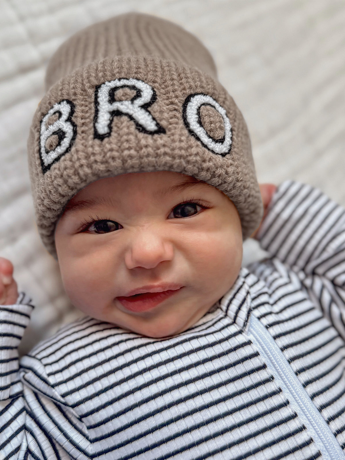 Baby wearing a knitted beige beanie with "BRO" written on it, smiling with a striped onesie.