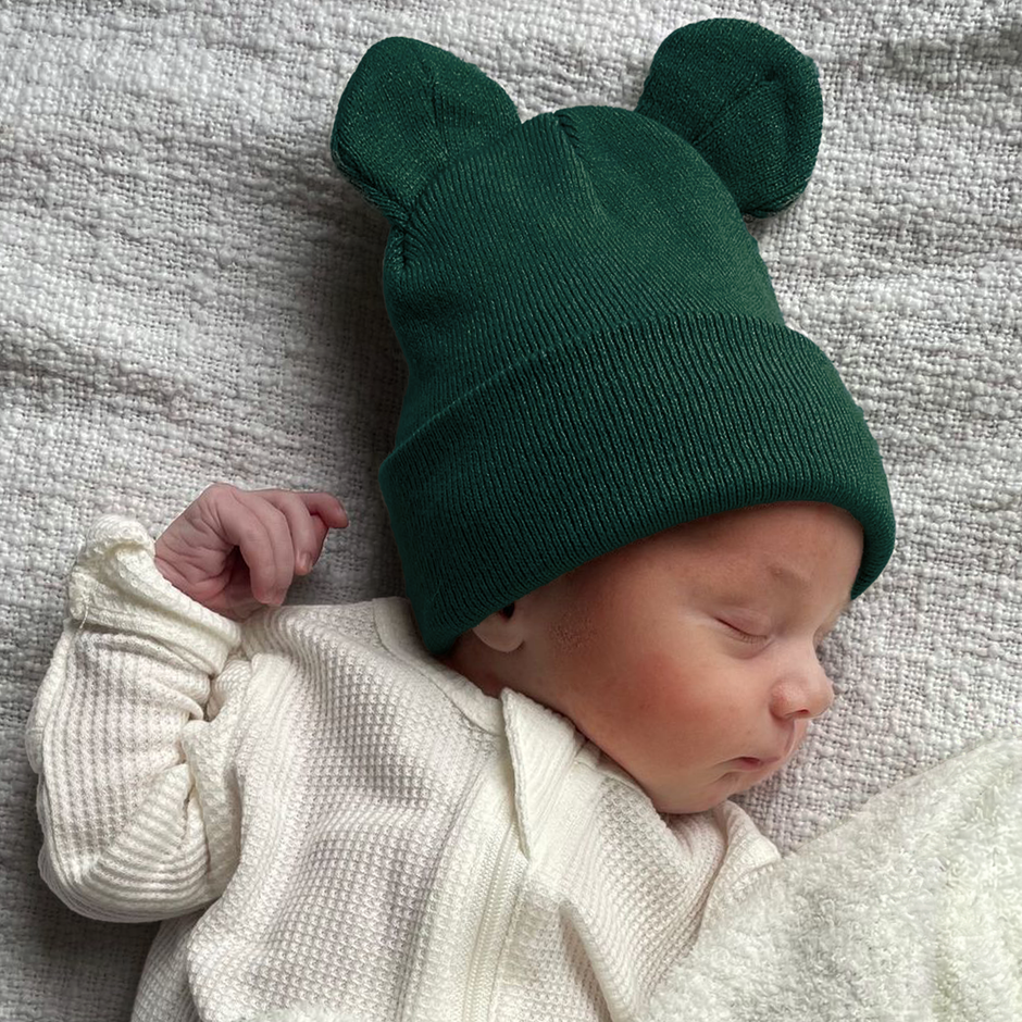 Sleeping baby in a green bear-ear beanie, wearing a white outfit, on a textured gray blanket.
