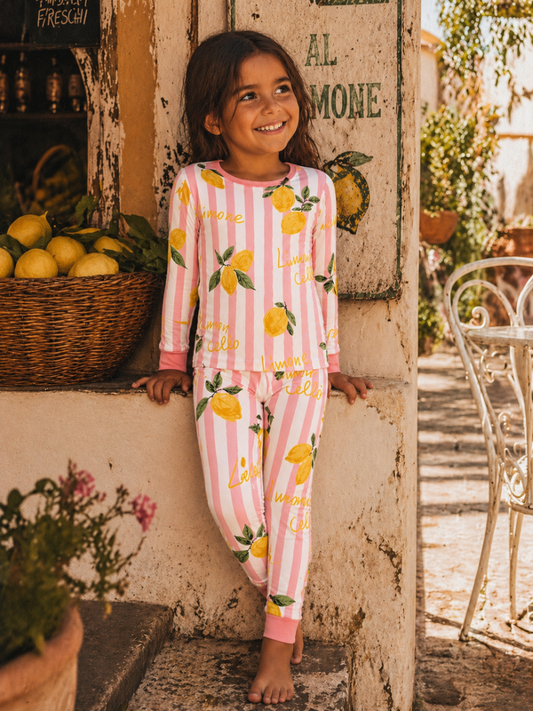 Smiling girl in lemon-patterned pajamas stands near a stone wall and lemon basket, enjoying a sunny day outdoors.