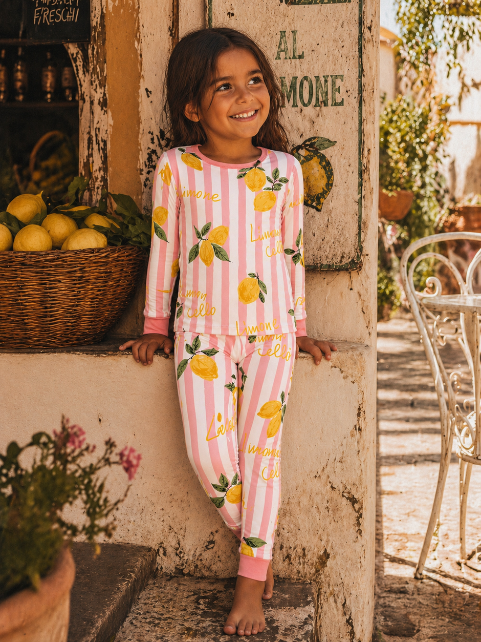 Smiling girl in lemon-patterned pajamas stands near a stone wall and lemon basket, enjoying a sunny day outdoors.