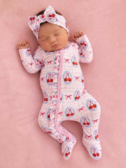 Newborn baby girl sleeping in a pink cherry-patterned onesie and matching headband on a soft pink blanket.