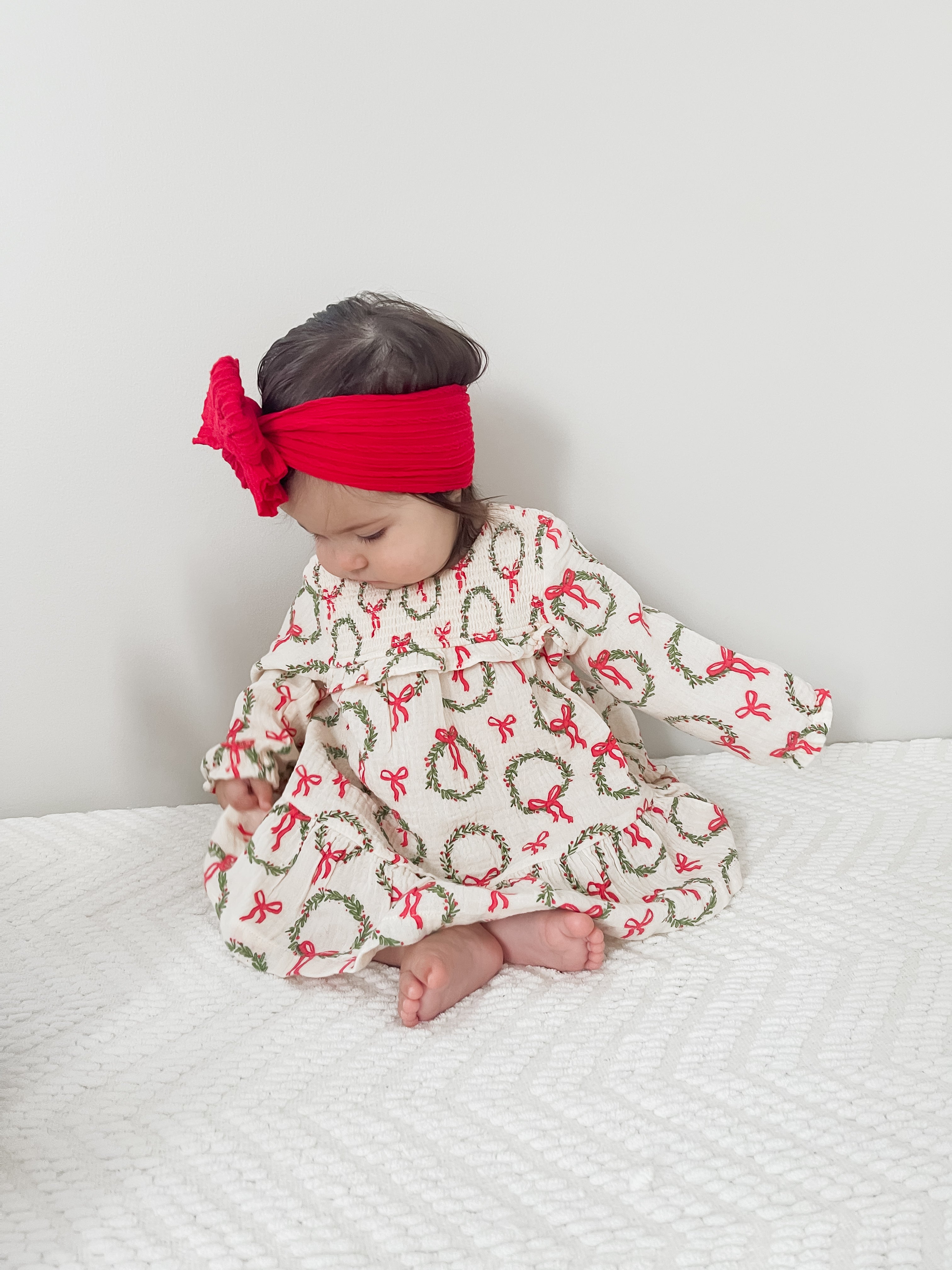 Baby wearing a festive dress with wreaths and bows, sitting on a bed, with a bright red headband.
