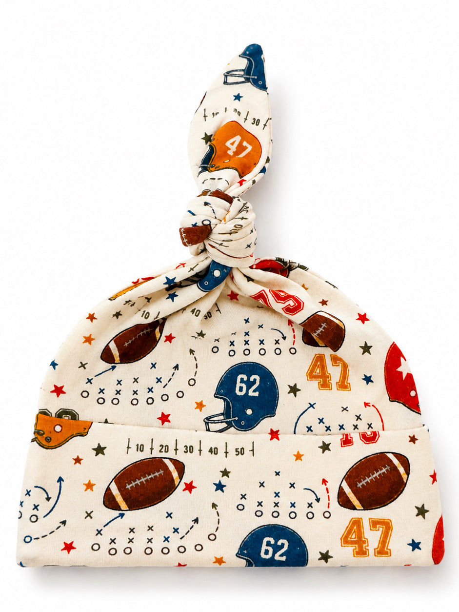 Football-themed baby hat with cute illustrations of helmets, footballs, and stars, tied at the top.
