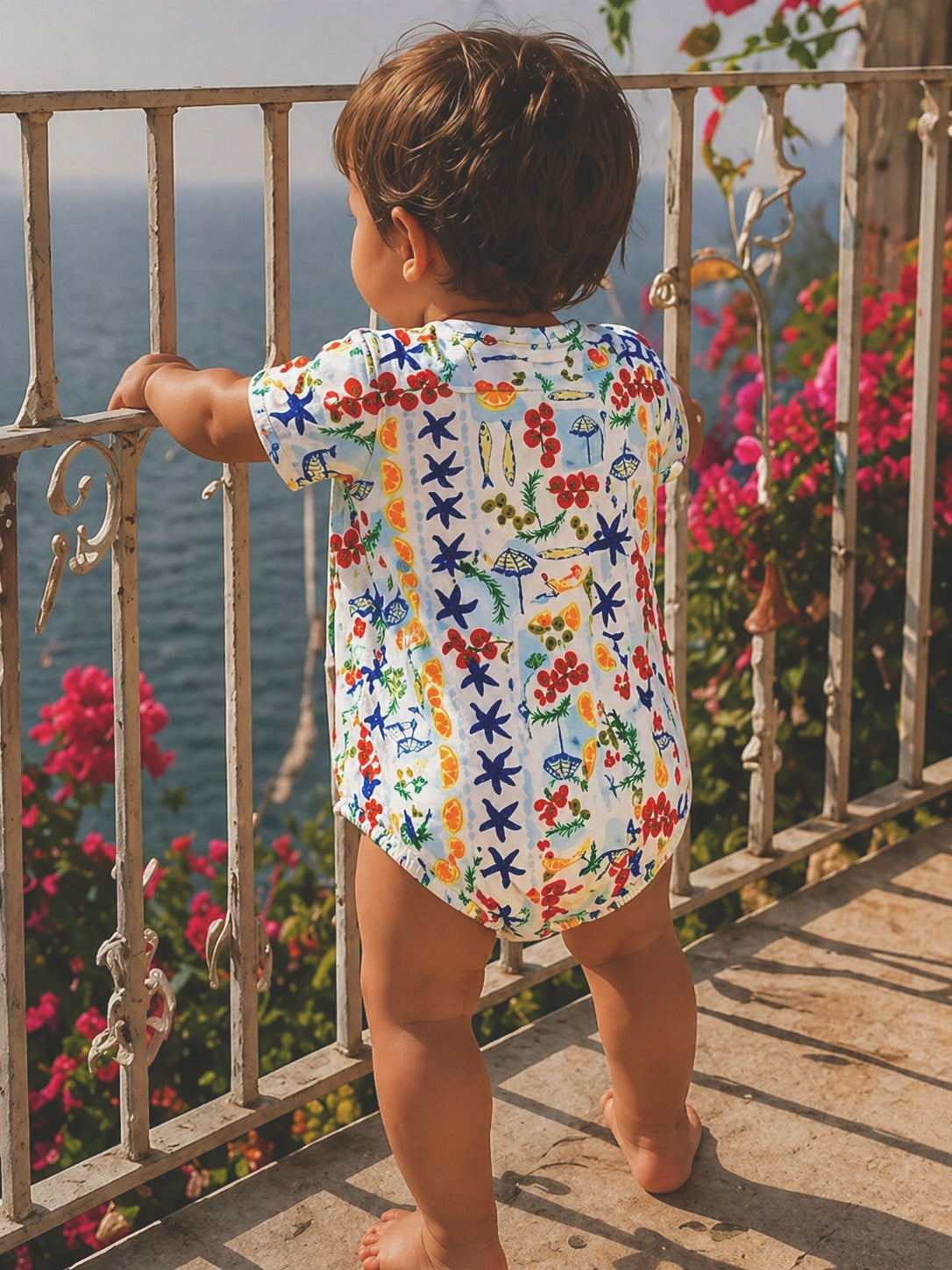 Child in colorful outfit gazes at the ocean, surrounded by flowers and a railing. A sunny day by the seaside.