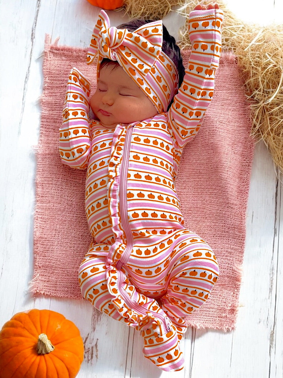 Baby girl asleep on pink blanket, wearing a pumpkin-patterned outfit and matching headband, surrounded by pumpkins.