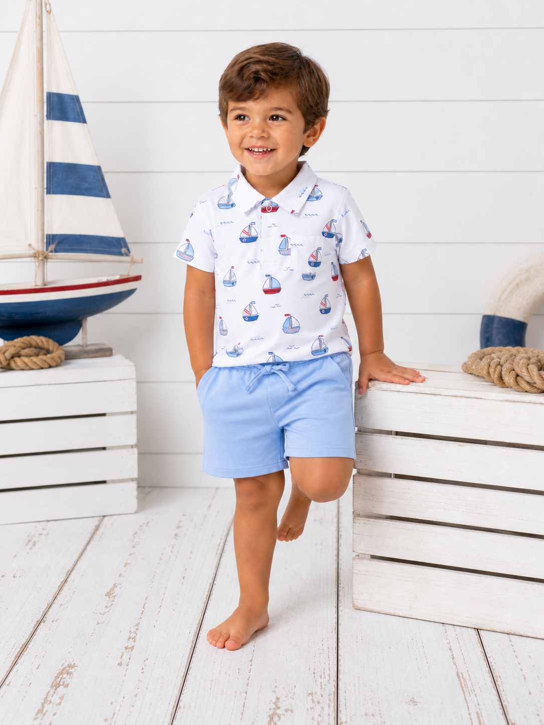 Smiling boy in a sailboat-themed shirt and blue shorts, standing next to a model sailboat and wooden crates.