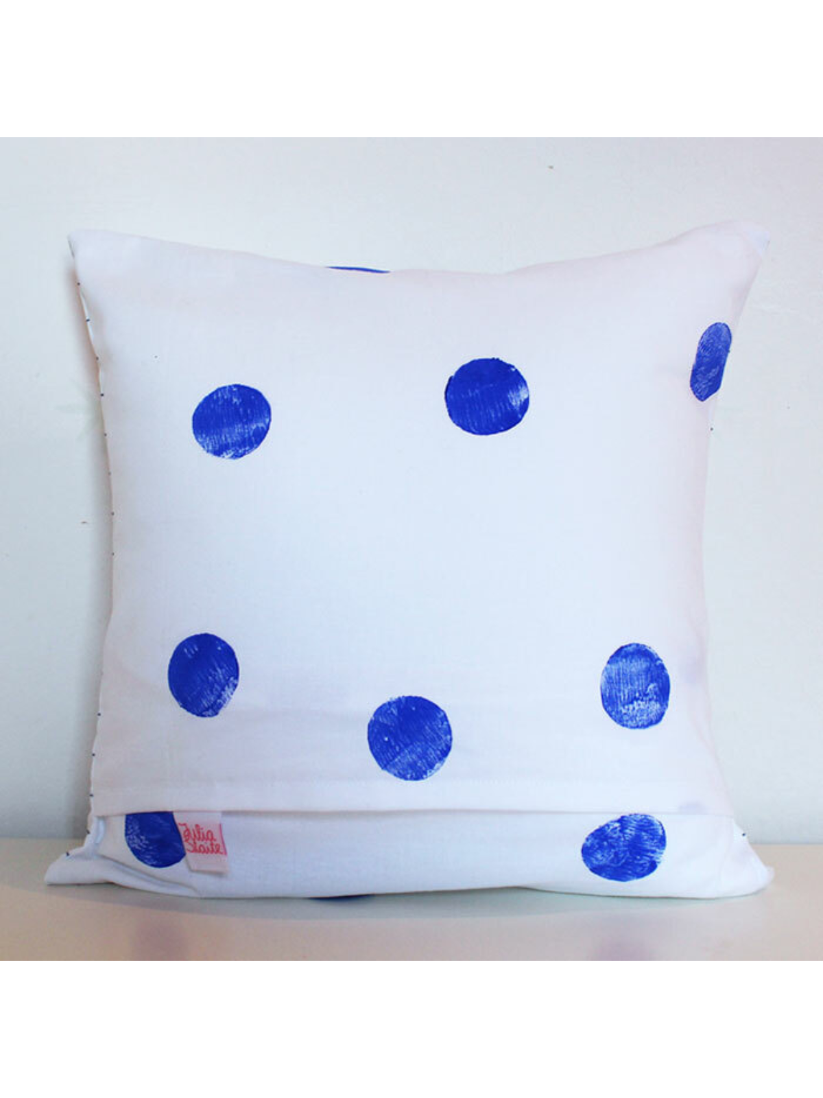 White pillow with blue polka dots against a light background, featuring a small brand label.
