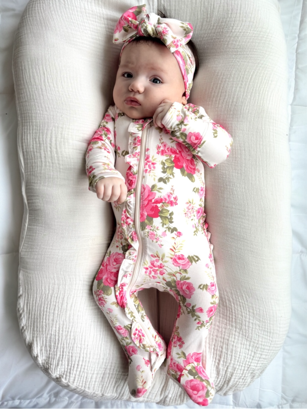Baby girl in floral onesie with a matching headband, lying on a soft, cream-colored surface.