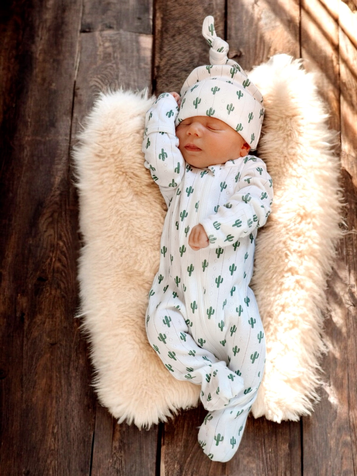 Sleeping newborn in cactus-patterned outfit on soft fur blanket, resting on a wooden surface.