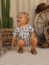 Smiling toddler in a patterned outfit squatting by cacti and rustic decor, capturing a joyful outdoor moment.