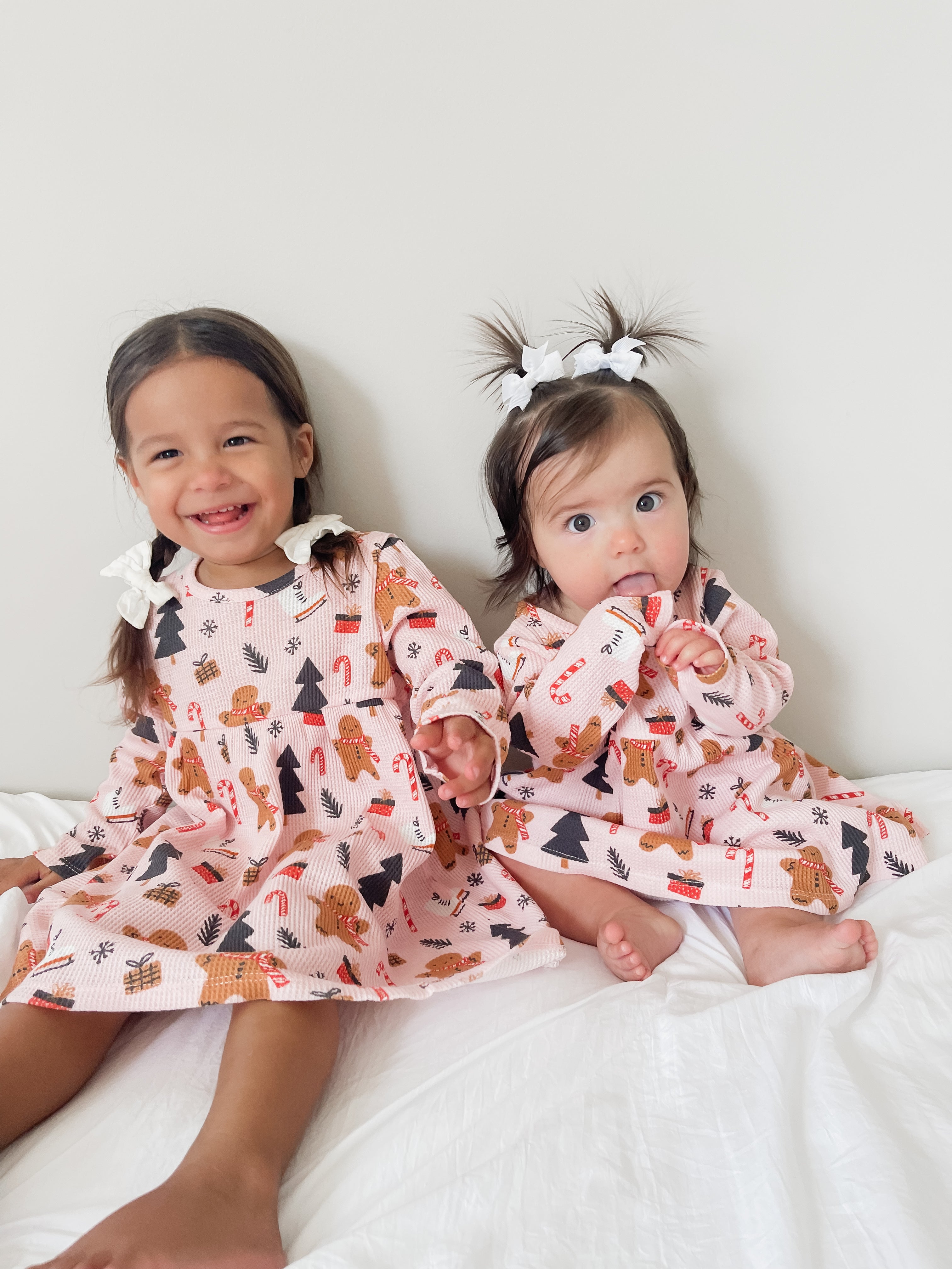 Two young girls in festive patterned dresses, smiling while sitting on a white bedspread against a neutral background.