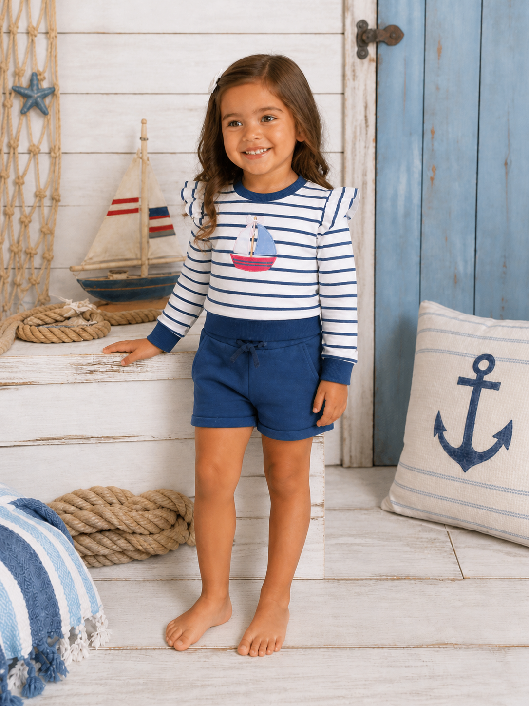 Smiling girl in a striped shirt with a sailboat graphic, navy shorts, standing in a nautical-themed setting.