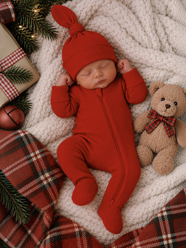 Newborn in a red outfit and hat, sleeping on a cozy blanket with a teddy bear and holiday decorations.
