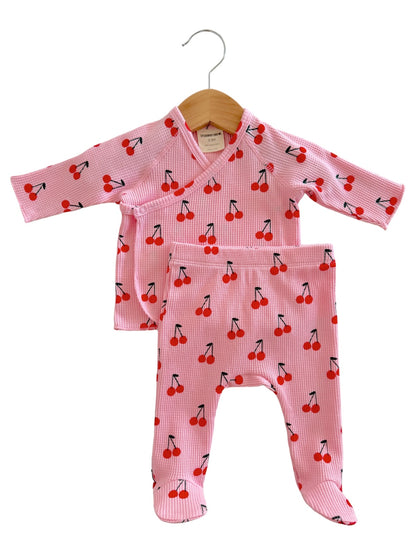 Pink baby outfit with cherry pattern, featuring a long-sleeve top and matching pants, hanging on a wooden hanger.