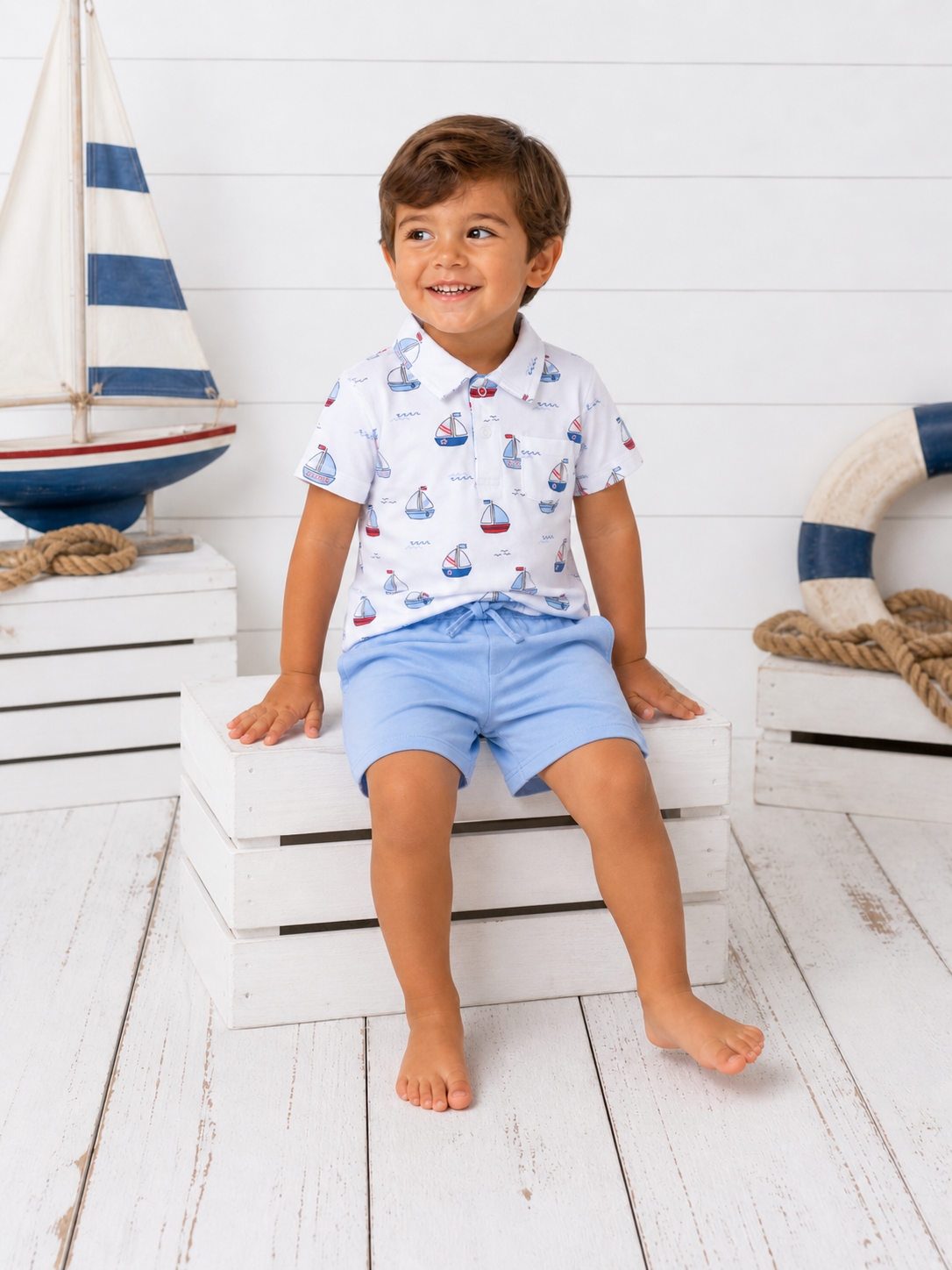 Smiling boy in a sailboat-printed shirt and blue shorts sitting on white crates by a nautical decor background.