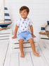 Smiling boy in a sailboat-printed shirt and blue shorts sitting on white crates by a nautical decor background.