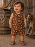 Smiling toddler in a brown patterned outfit stands near a wooden horse and barrels, set in a rustic outdoor scene.