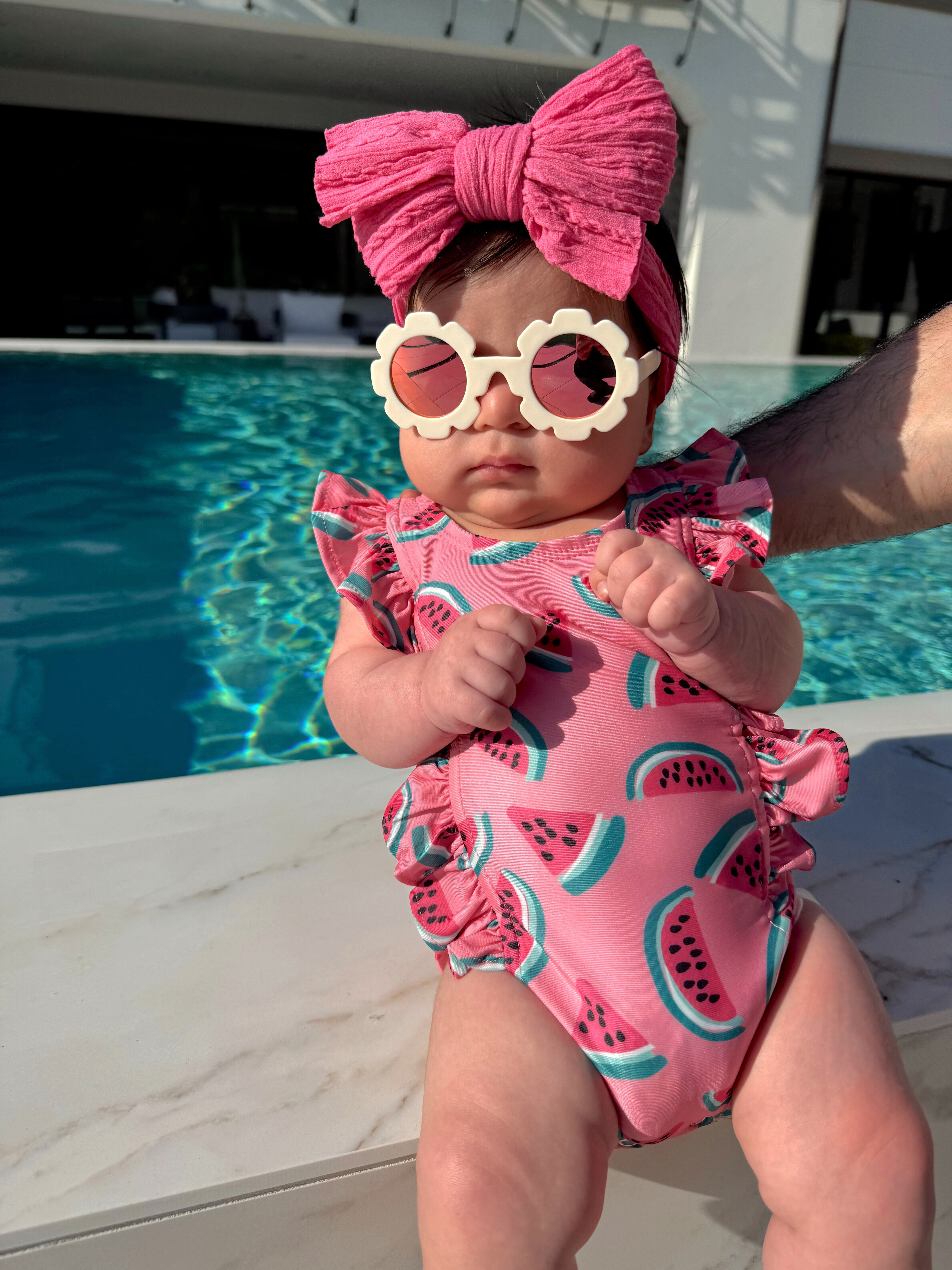 Baby in a pink watermelon swimsuit and big bow, wearing sunglasses by a pool on a sunny day.