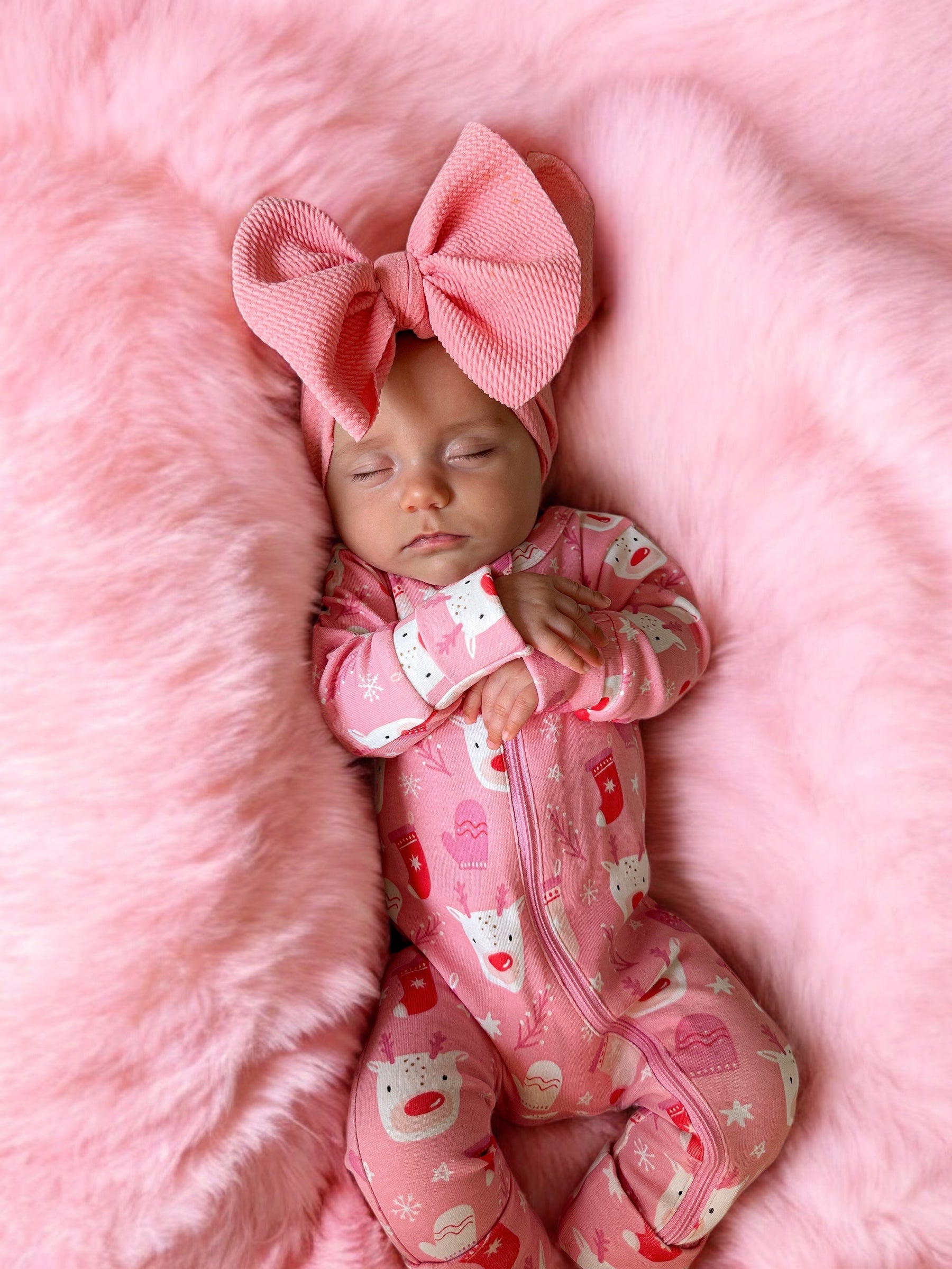 Sleeping baby in pink pajamas with a large pink bow on a fluffy pink blanket.