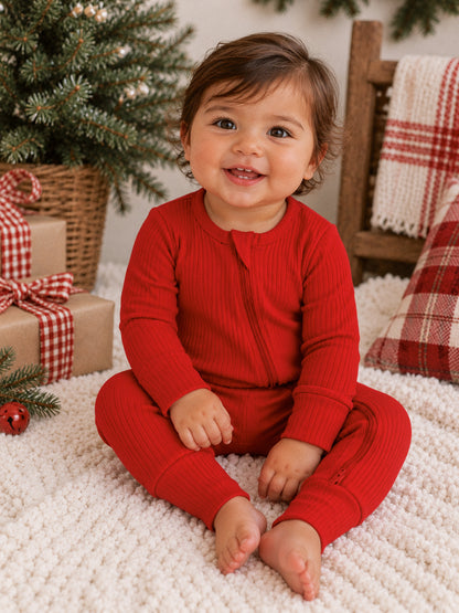 Smiling toddler in a red knit onesie sits on a cozy blanket with gifts and holiday decor in the background.