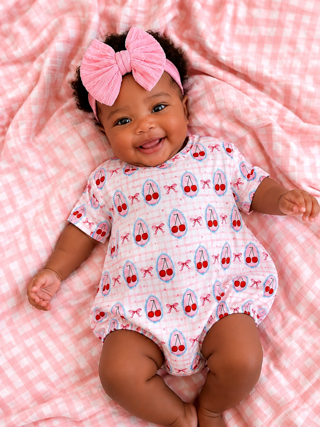 Smiling baby in a cherry-patterned onesie and pink bow, lying on a pink checkered blanket.