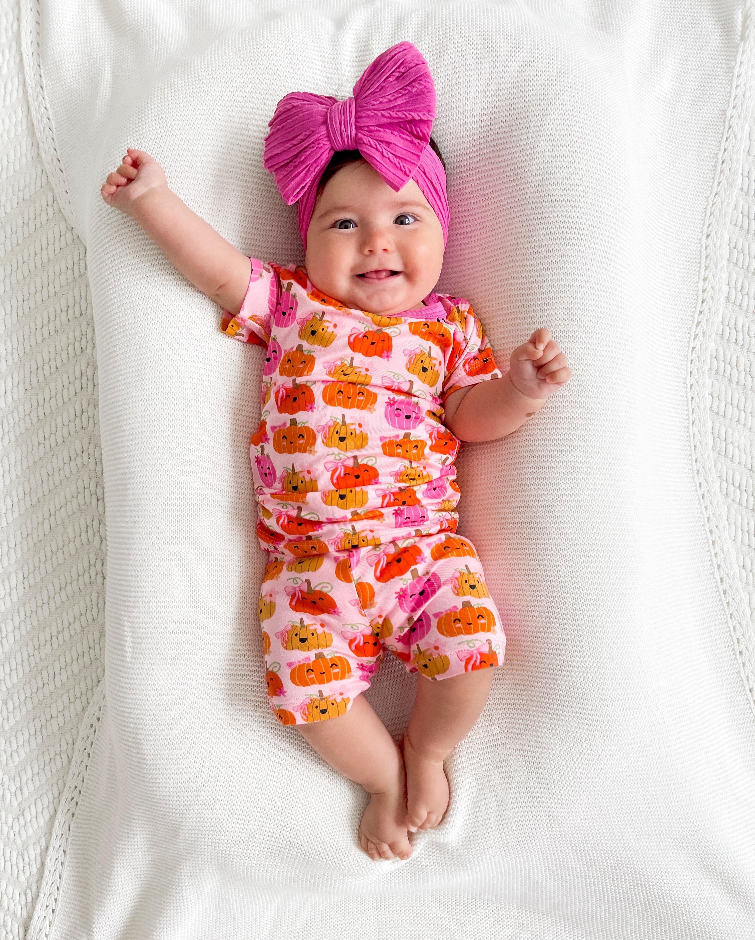 Smiling baby in a pumpkin-patterned outfit with a large pink bow, lying on a soft white blanket.