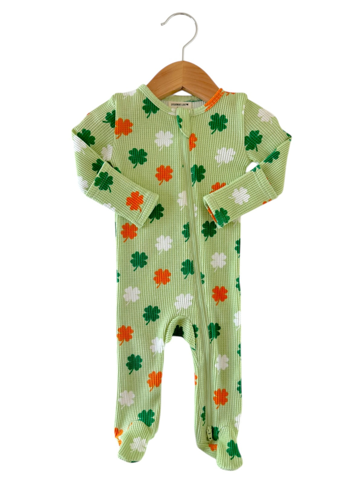 Green infant onesie with shamrock pattern in orange and white, hanging on a wooden hanger against a white background.