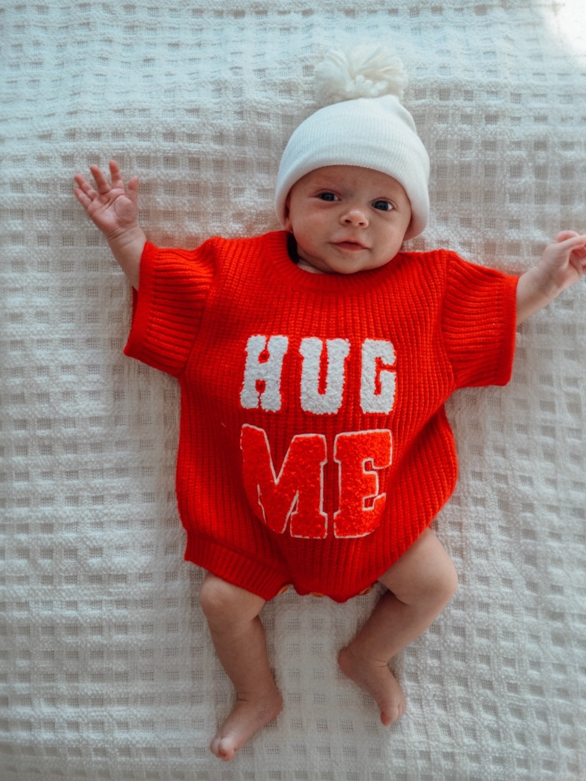 Baby wearing a red sweater with "HUG ME" text, lying on a textured white blanket.