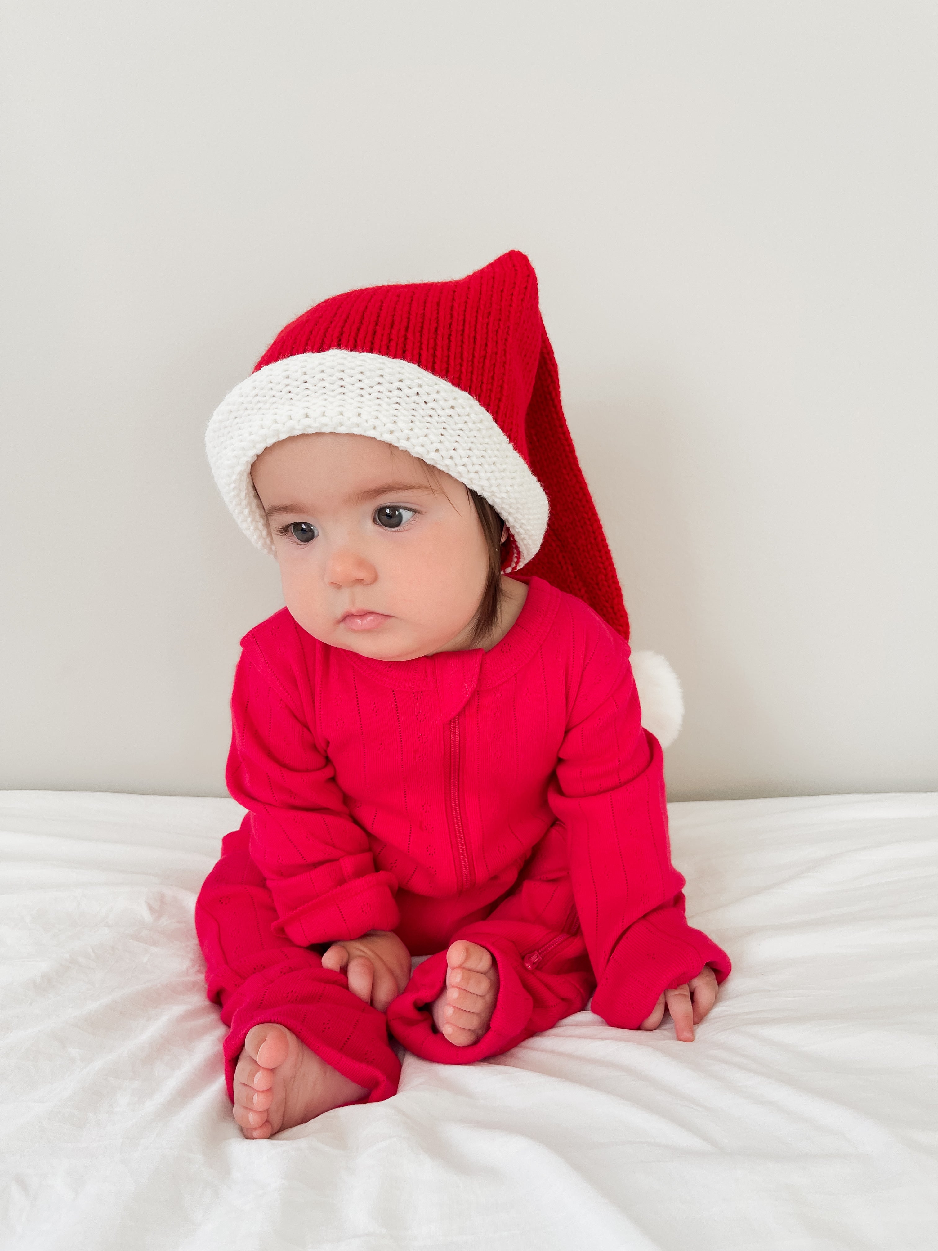 Baby in a red outfit with a white-trimmed Santa hat, sitting on a white surface against a light background.