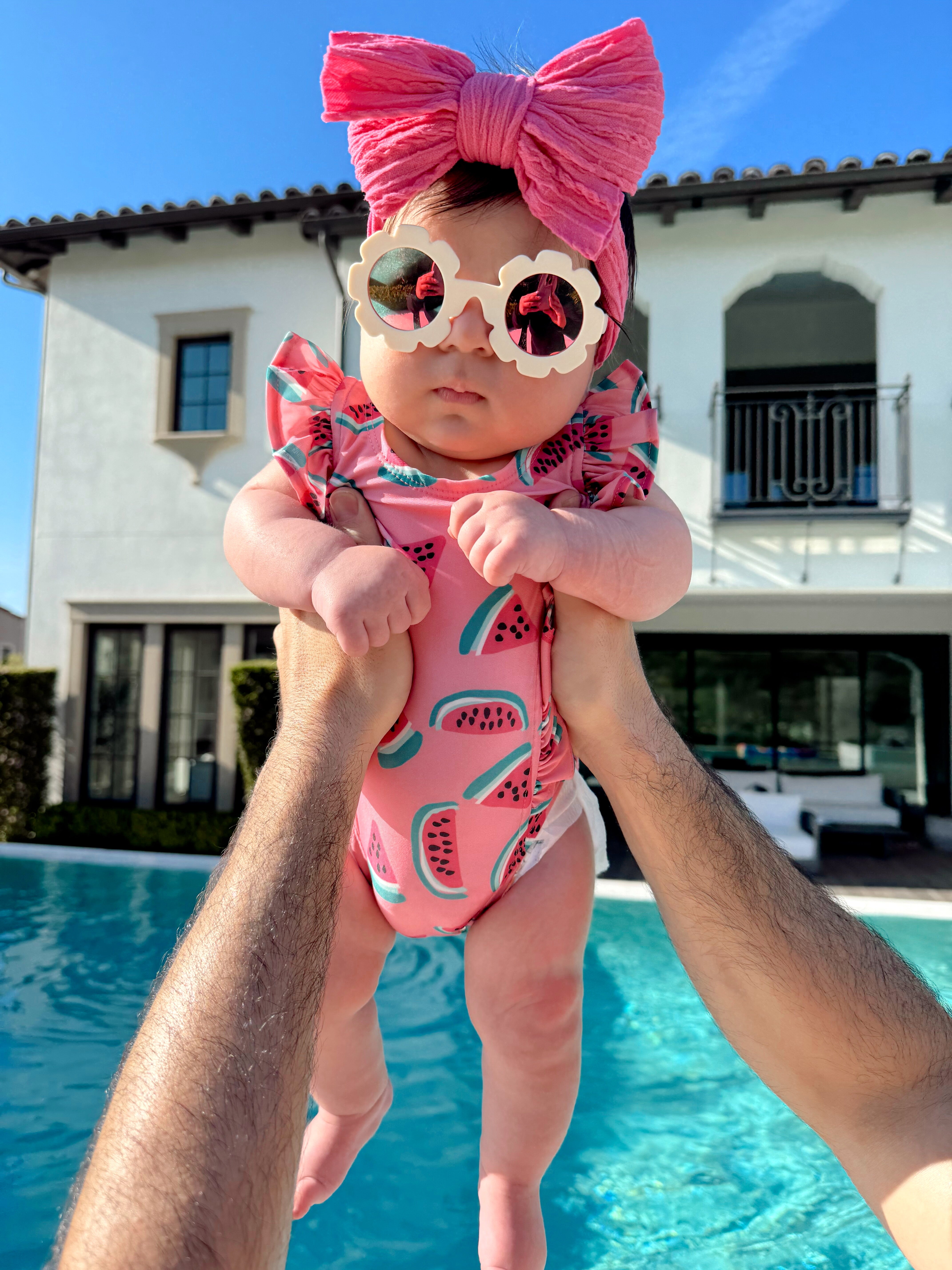Baby in a watermelon swimsuit and pink bow sunglasses held above a poolside, with a sunny backdrop of a villa.
