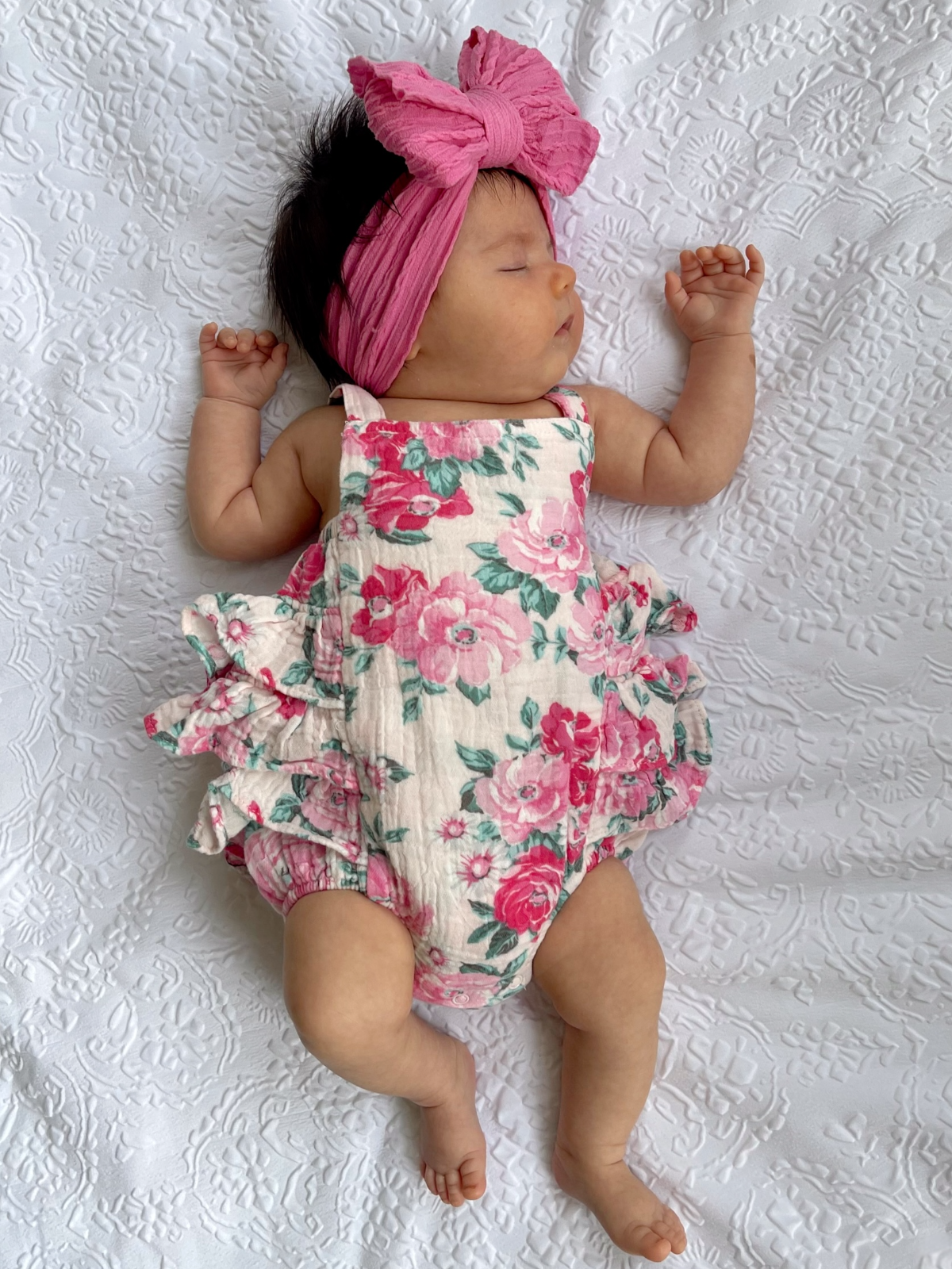 Baby girl in a floral romper and pink headband, peacefully sleeping on a white textured blanket.