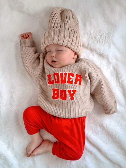 Sleeping baby in beige sweater and red pants, wearing a cozy knit hat with "LOVER BOY" text, on a soft white blanket.