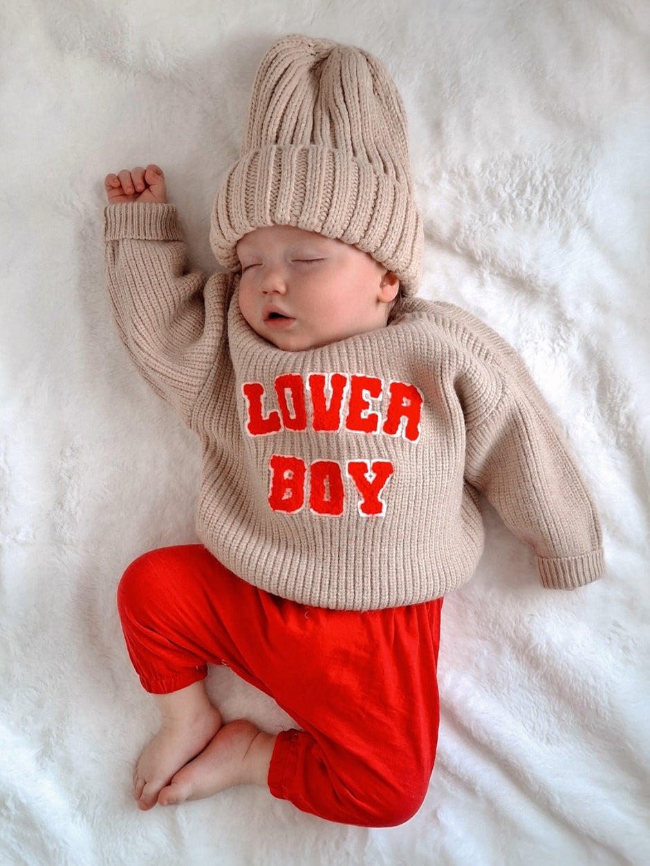 Sleeping baby in beige sweater and red pants, wearing a cozy knit hat with "LOVER BOY" text, on a soft white blanket.