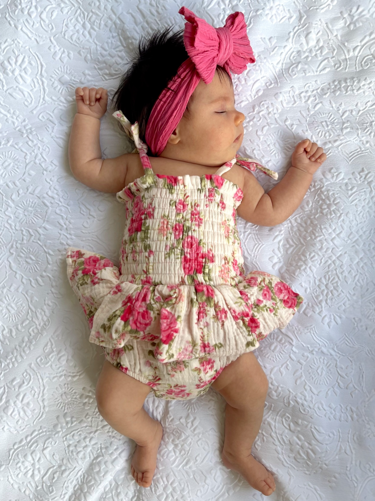 Baby girl in a floral outfit and pink bow headband, peacefully sleeping on a white textured blanket.