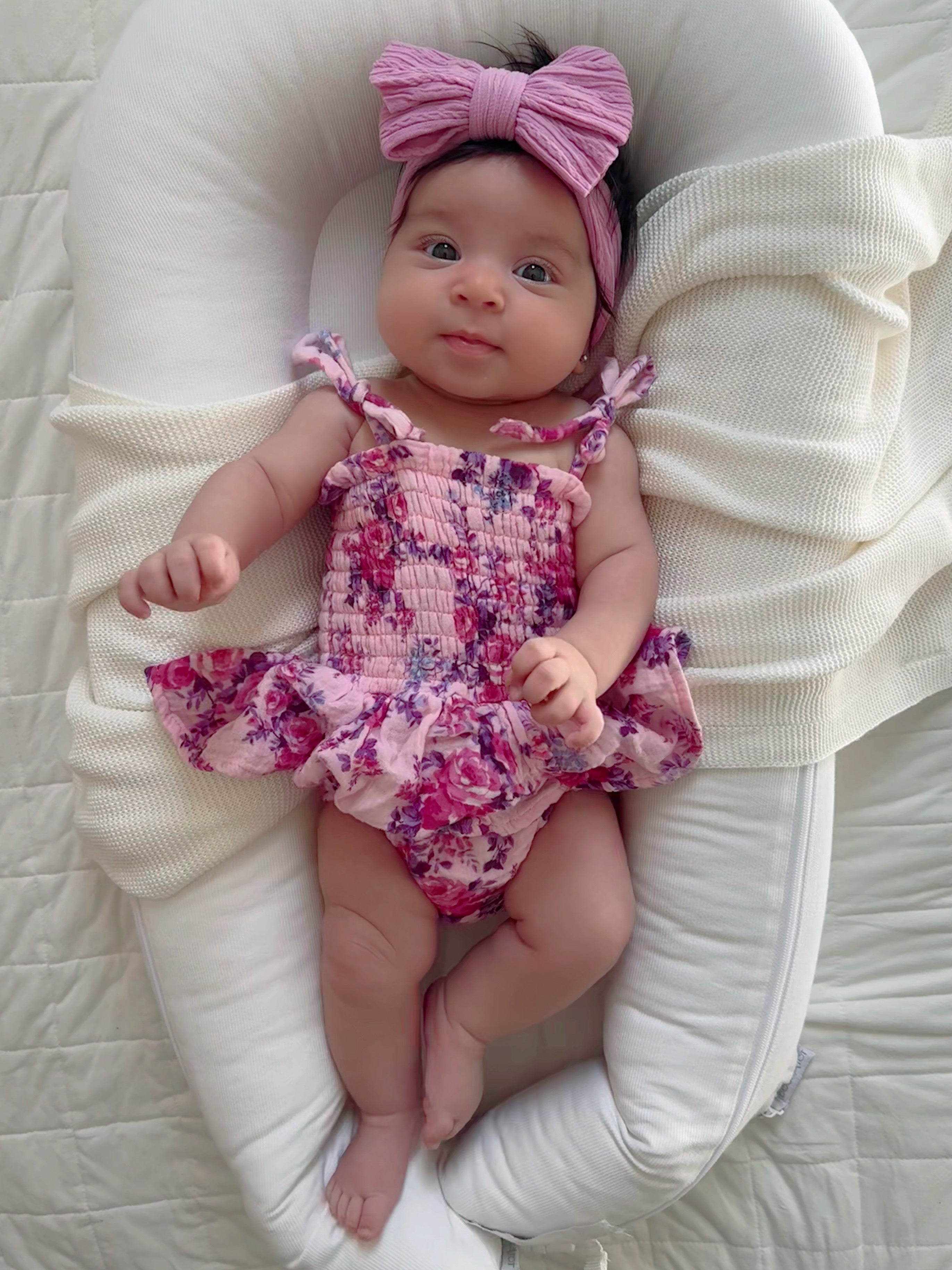 Smiling baby girl in a floral swimsuit and pink bow, lying on a soft, cushioned surface.