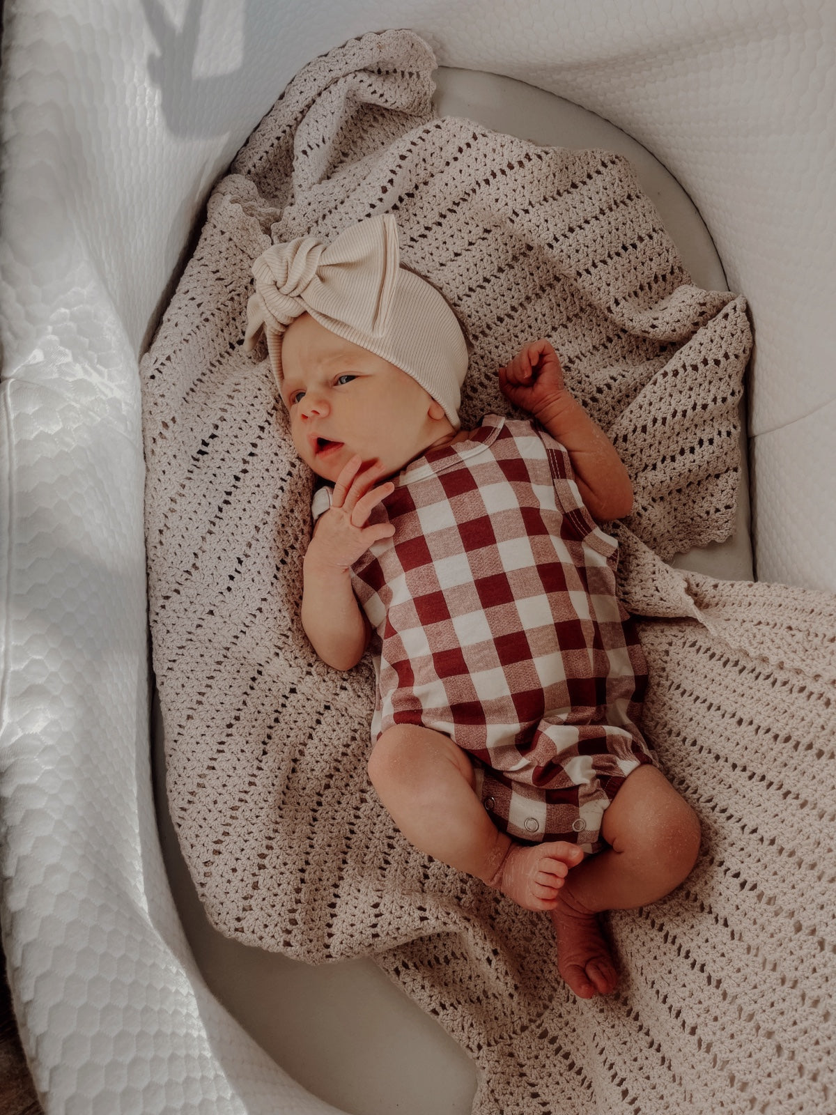 Infant lying on a textured blanket, wearing a checkered romper and a bow headband. Soft, warm lighting.