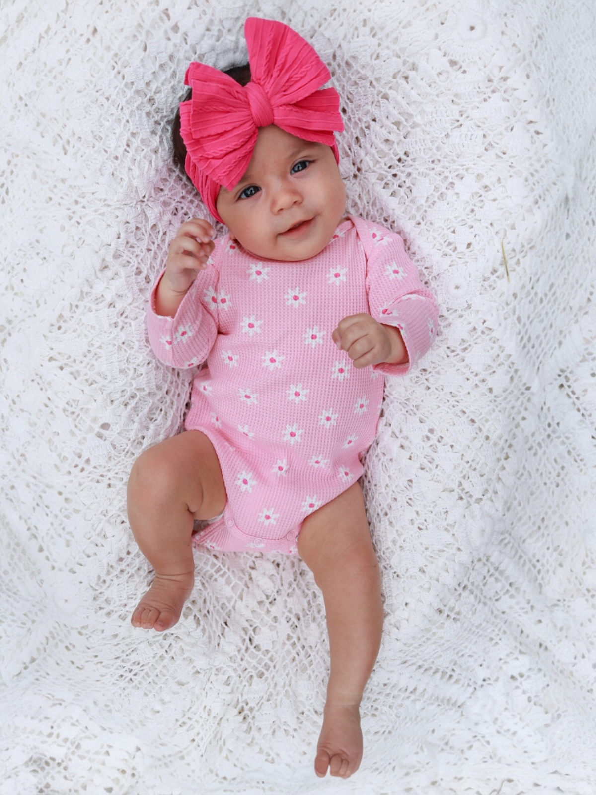 Infant girl in a pink floral onesie and large pink bow, lying on a textured white blanket.