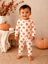 Smiling toddler in pumpkin-patterned onesie standing on a textured surface with pumpkins in the background.