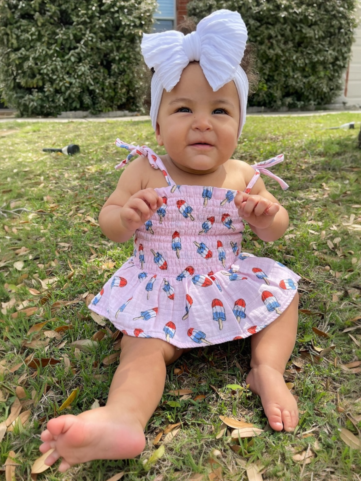 Smiling infant sits on grass wearing a pink dress with popsicle prints and a large white bow headband.