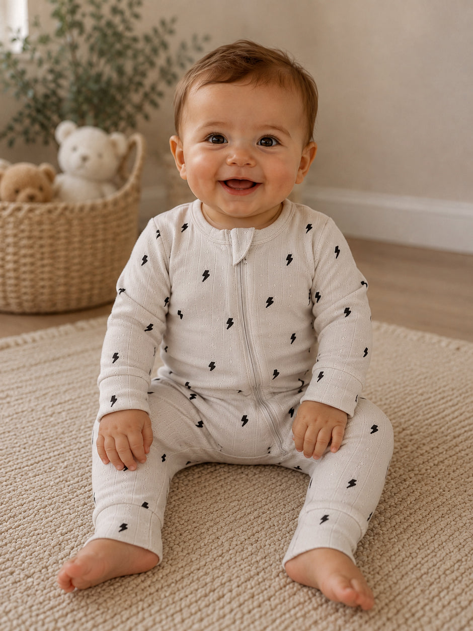 Smiling baby in a light onesie with lightning patterns, sitting on a rug with teddy bears in the background.