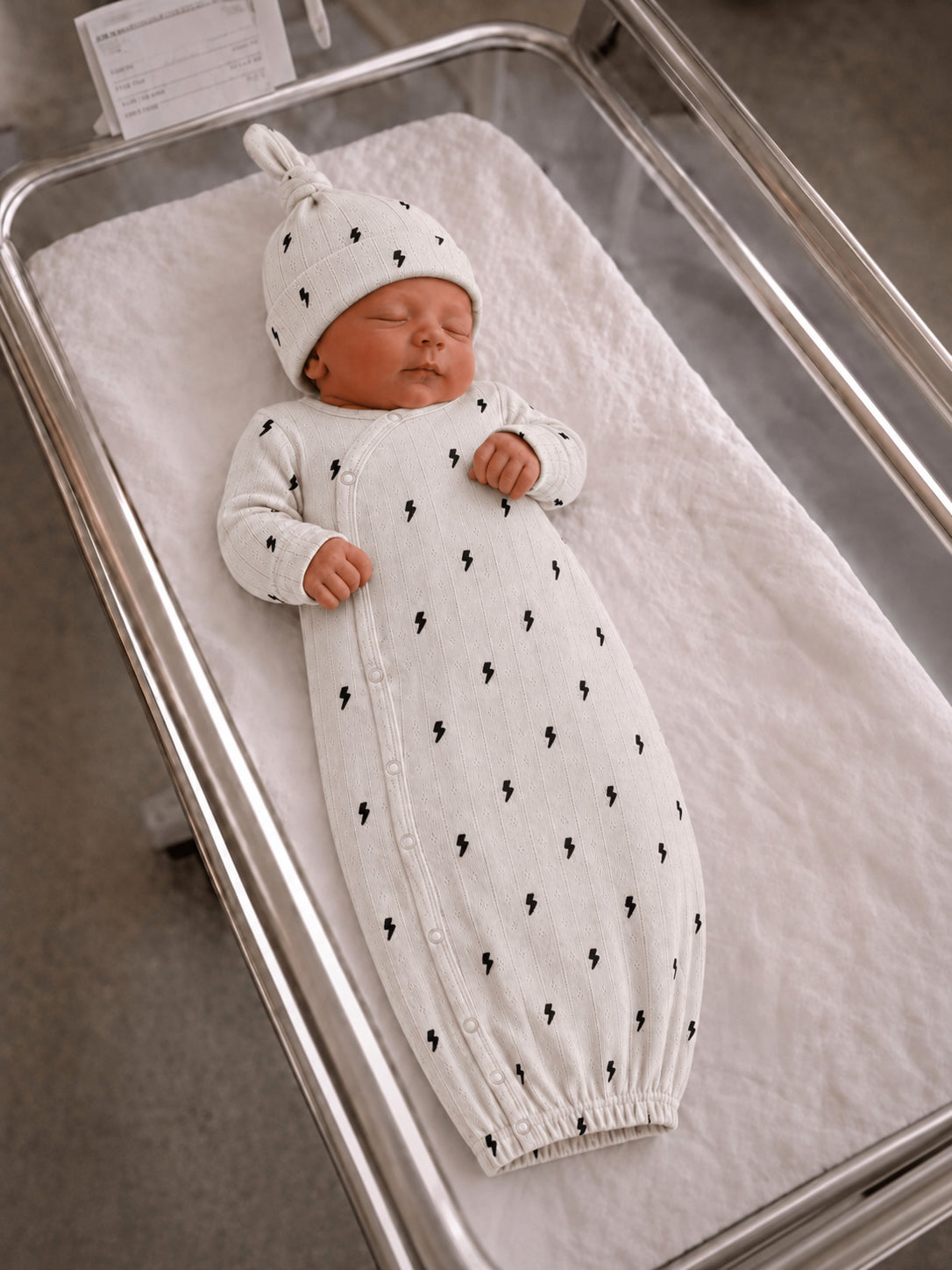 Newborn asleep in a cozy white outfit with lightning bolt patterns, resting in a hospital bassinet.