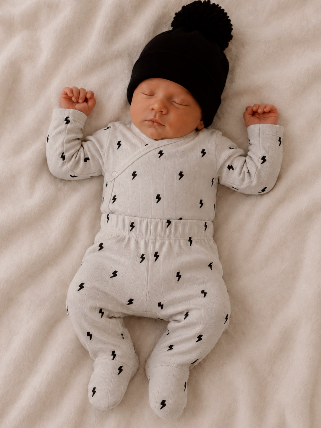 Newborn baby peacefully sleeping in gray outfit with lightning patterns and black hat on a soft blanket.
