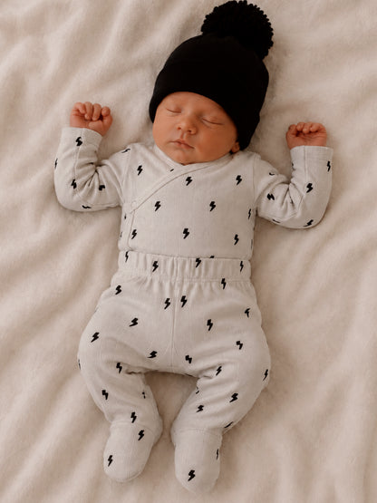 Newborn baby peacefully sleeping in gray outfit with lightning patterns and black hat on a soft blanket.