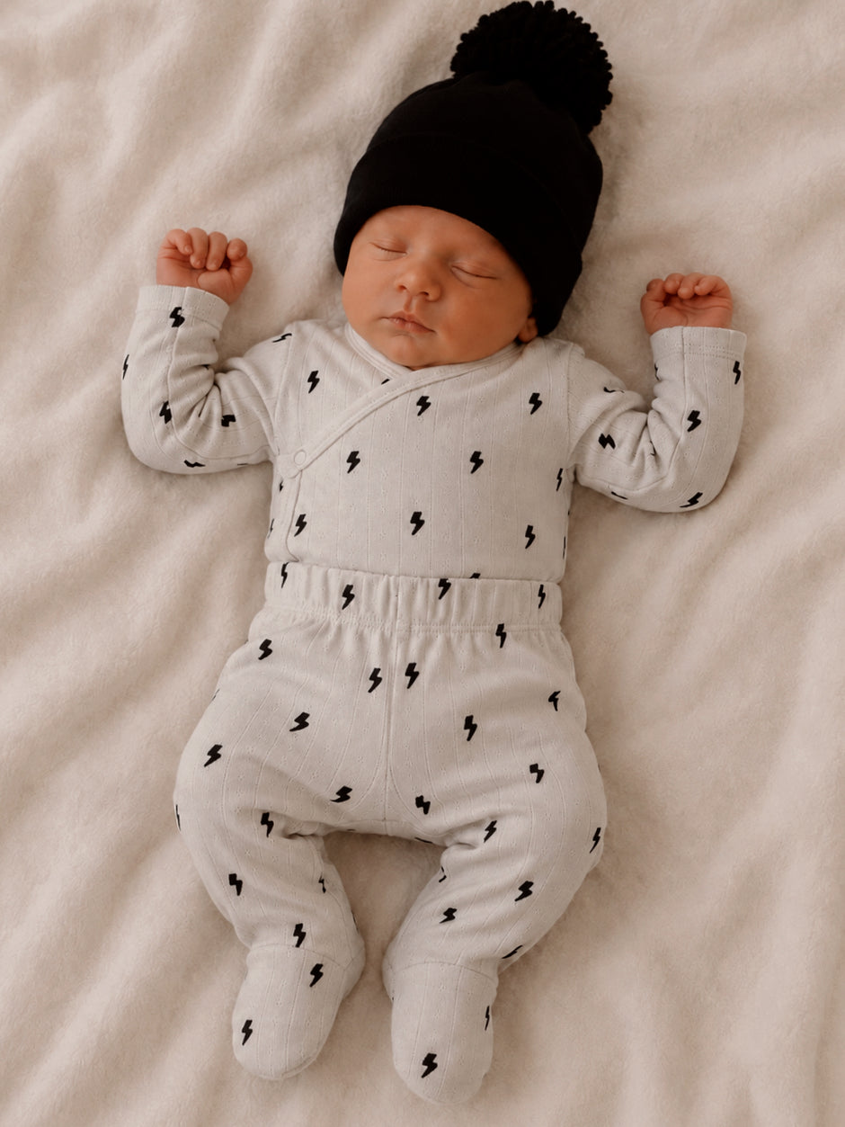 Newborn baby peacefully sleeping in gray outfit with lightning patterns and black hat on a soft blanket.