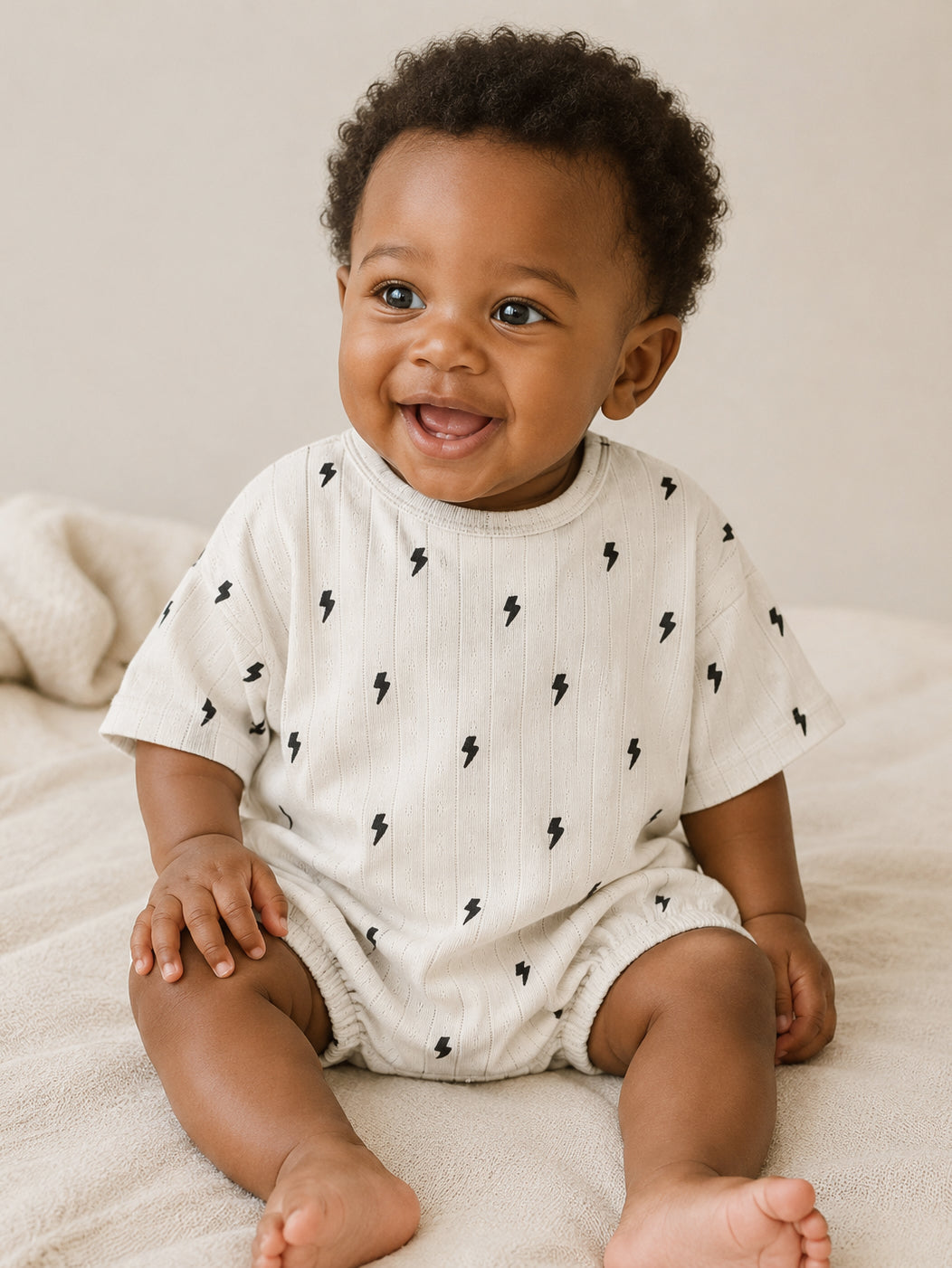 Smiling baby in a light outfit with lightning bolt patterns, sitting on a soft surface.